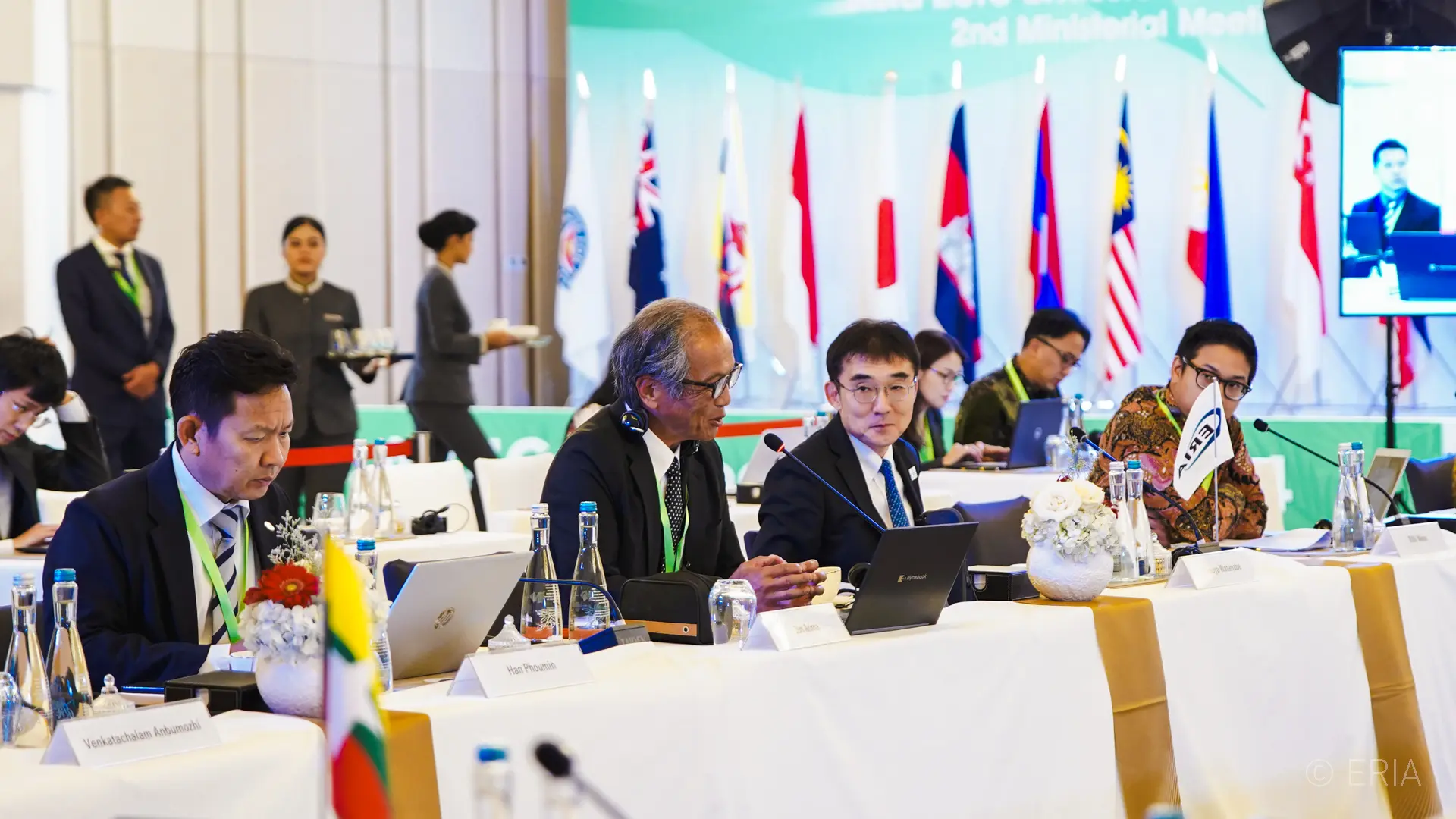A diverse group of individuals in formal attire participate in a conference. Several international flags are displayed behind them, conveying a diplomatic atmosphere.
