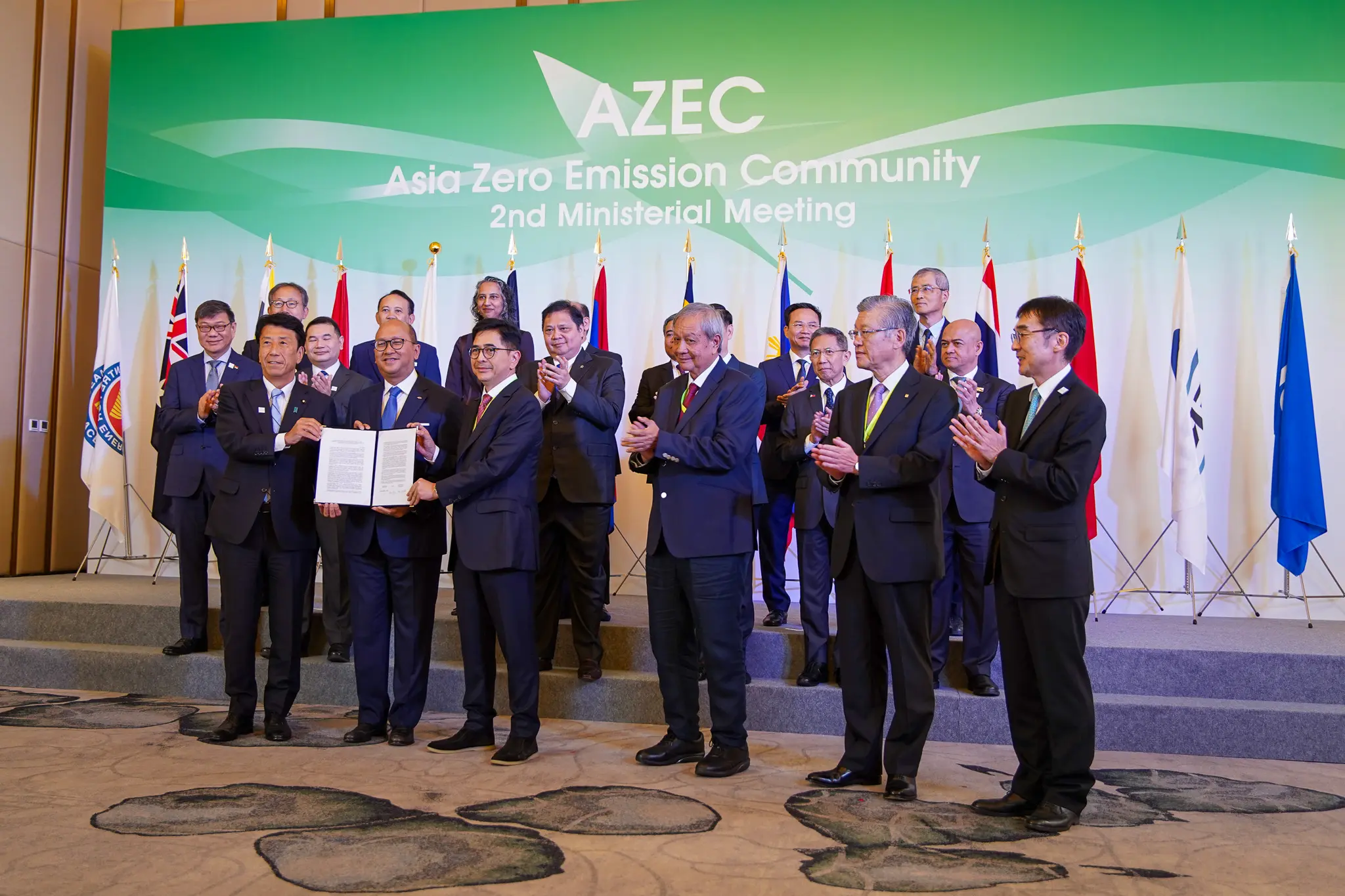 A group of officials stands smiling onstage at the Asia Zero Emission Community 2nd Ministerial Meeting. They hold a document, surrounded by various national flags.